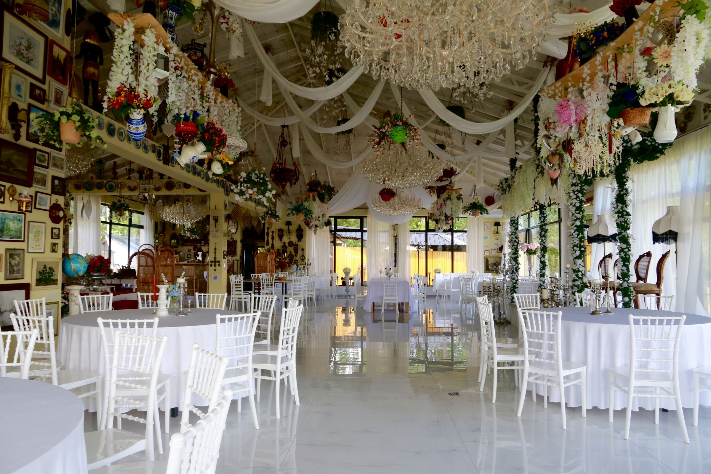 The dining room laid for a celebration — round tables, white chairs, chandeliers and garlands beneath the high ceiling