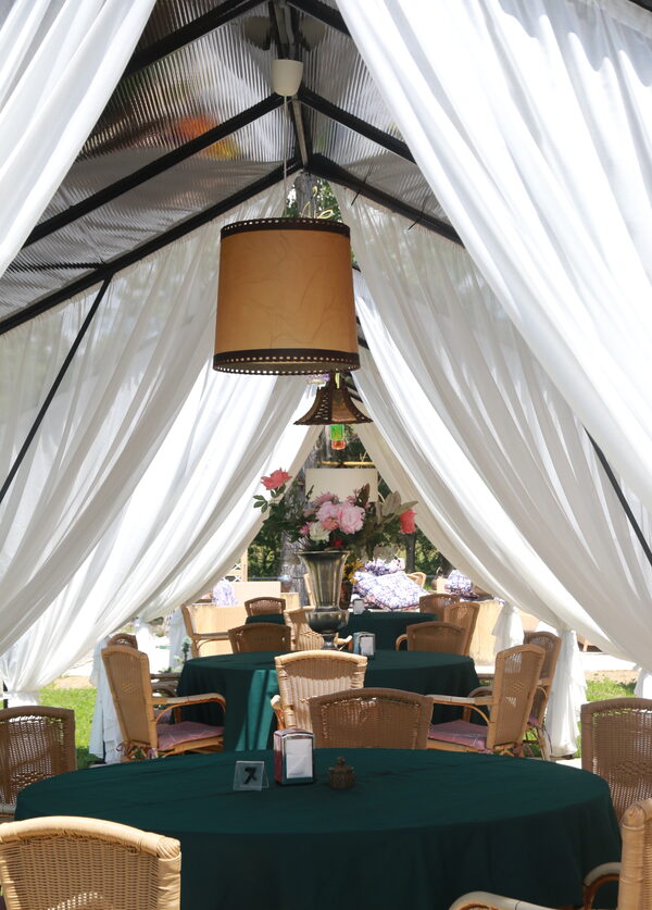 Inside a tent, a paper lampshade hanging above green-clothed tables and wicker chairs